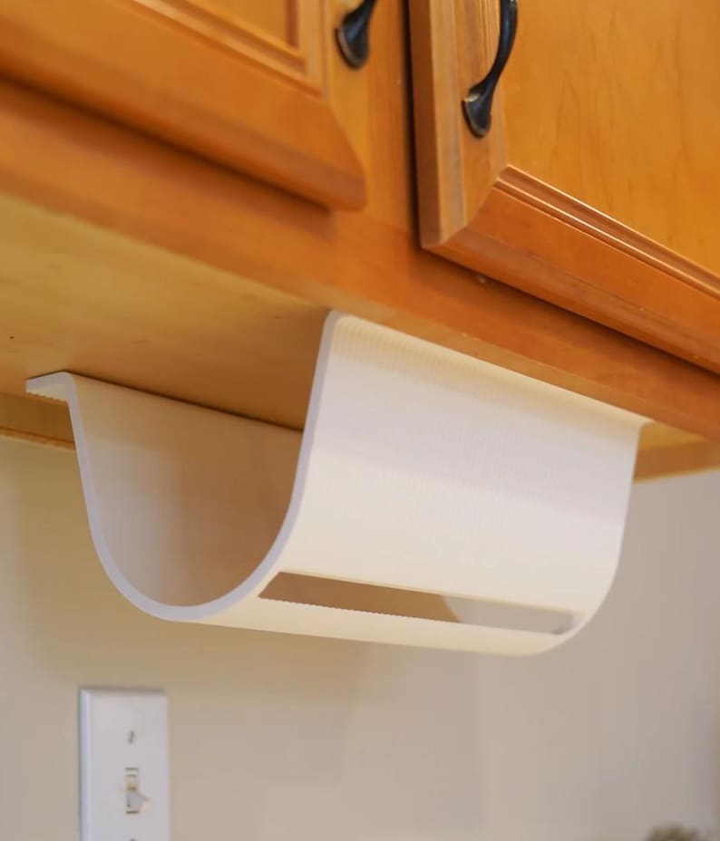 May include: A white, curved paper towel holder mounted under a wooden cabinet. The holder has a rectangular opening and is designed to dispense paper towels. The cabinet has dark handles.