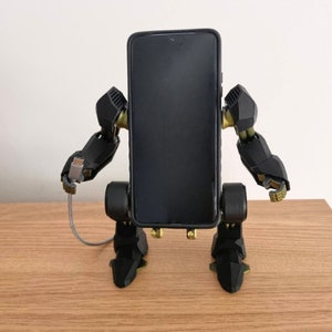 May include: A black phone stand shaped like a robot with gold accents. The stand has a black phone in it and a charging cable plugged into the phone.