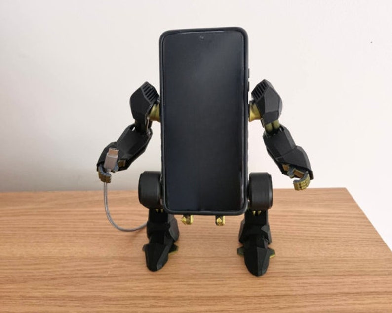 May include: A black phone stand shaped like a robot with gold accents. The stand has a black phone in it and a charging cable plugged into the phone.
