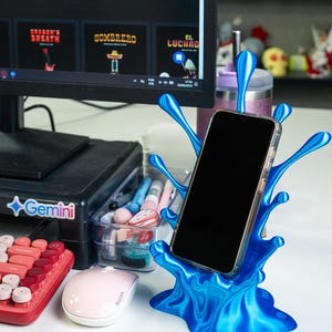 May include: A blue 3D printed phone stand shaped like a water splash. The stand is holding a black smartphone.