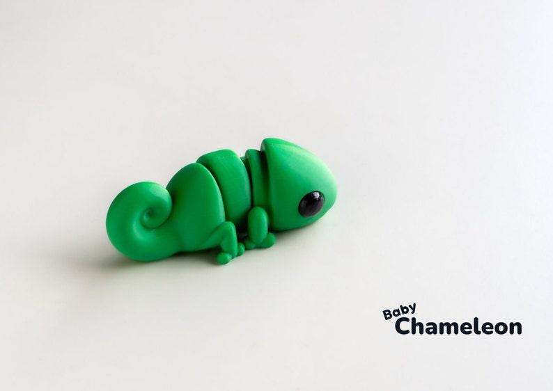 May include: A small green chameleon figurine with black eyes. The figurine is made of a smooth, pliable material and has a curled tail. The text "Baby Chameleon" is printed below the figurine.