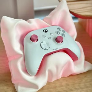 Könnte beinhalten: Ein weißer und rosa Gamecontroller mit rosa Knöpfen und einem rosa Akzent an der Unterseite. Der Controller liegt auf einem rosa Kissen.