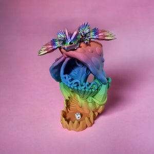 May include: A rainbow-colored dice tower with a dragon figurine on top. The tower is made of 3D printed plastic and has a compartment for dice at the bottom.