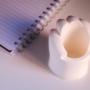 May include: White ceramic hand-shaped planter with a pink accent on the fingertip. The planter is empty and ready for a small plant.