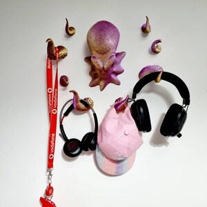 May include: A pink and gold octopus wall decor with eight tentacles, a red lanyard with the Vodafone logo, a pair of black headphones, and a pink and white baseball cap.