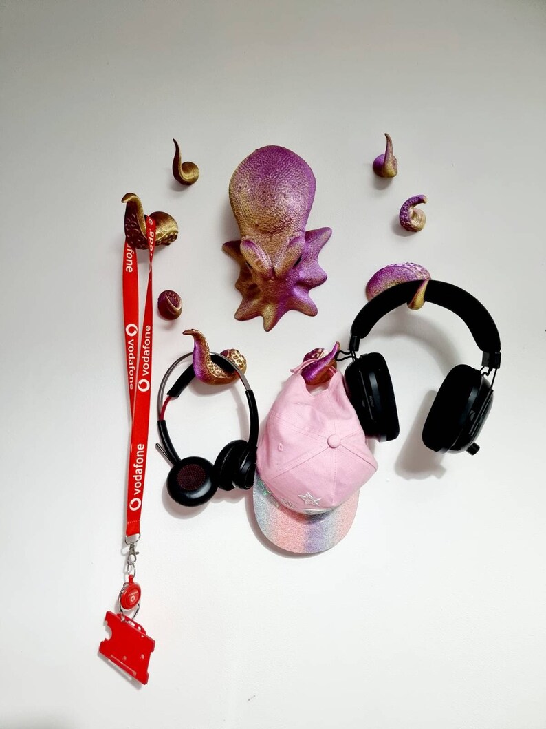 May include: A pink and gold octopus wall decor with eight tentacles, a red lanyard with the Vodafone logo, a pair of black headphones, and a pink and white baseball cap.