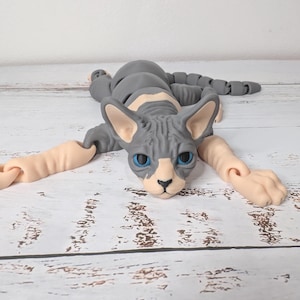 May include: A gray and white Sphynx cat figurine with articulated limbs. The cat has blue eyes and is lying down with its arms and legs extended. The figurine is made of a smooth material and rests on a wooden surface.