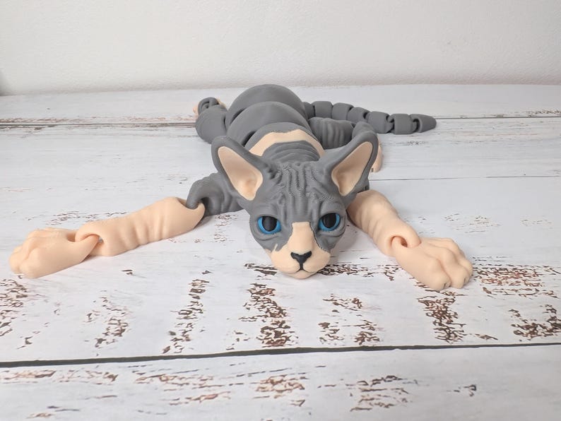 May include: A gray and white Sphynx cat figurine with articulated limbs. The cat has blue eyes and is lying down with its arms and legs extended. The figurine is made of a smooth material and rests on a wooden surface.
