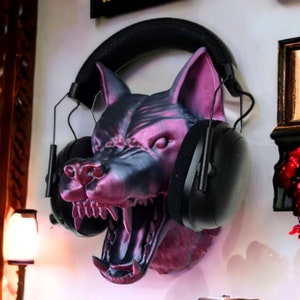 May include: A wall-mounted headphone holder shaped like a snarling wolf's head. The sculpture is a mix of black and purple, with the headphones resting on top. The wolf's mouth is open, revealing sharp teeth.