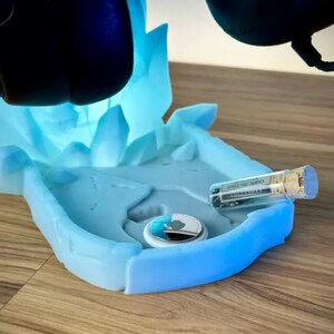 May include: A blue and white 3D printed stand with a frosted finish. The stand has a circular indentation for a small object and a small glass vial with a cork stopper. The stand is designed to hold a pair of headphones.