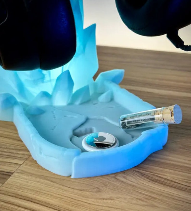 May include: A blue and white 3D printed stand with a frosted finish. The stand has a circular indentation for a small object and a small glass vial with a cork stopper. The stand is designed to hold a pair of headphones.