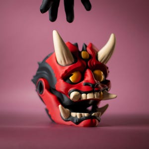 May include: A red and black Oni mask with white horns and teeth. The mask has yellow eyes and is set against a pink background. A black gloved hand hovers above the mask.