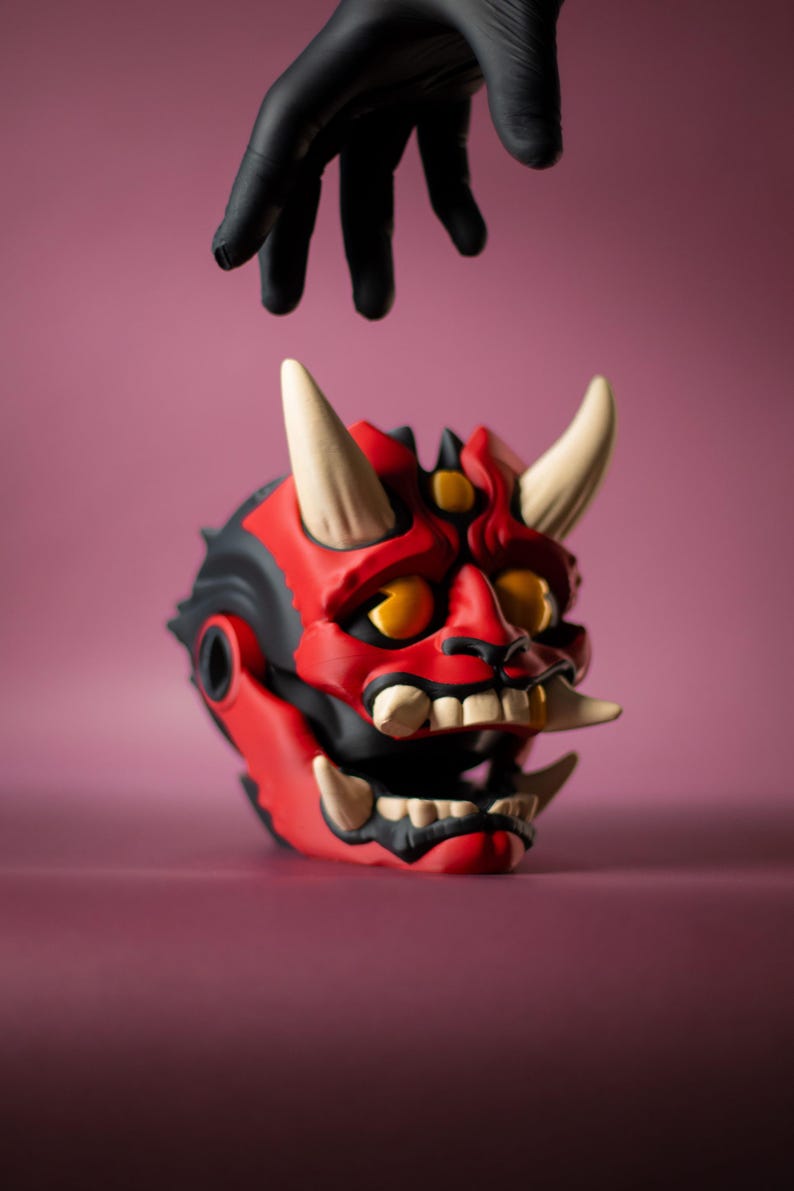 May include: A red and black Oni mask with white horns and teeth. The mask has yellow eyes and is set against a pink background. A black gloved hand hovers above the mask.