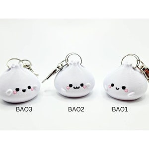 May include: Three white, cartoon-style,  bao bun keychains with smiling faces. Each keychain has a silver ring and a silver clasp.