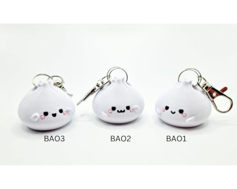 May include: Three white, cartoon-style,  bao bun keychains with smiling faces. Each keychain has a silver ring and a silver clasp.