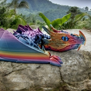 May include: A colourful, rainbow-hued dragon sculpture with a blue eye, lying on a grey rock. The dragon is made of 3D printed plastic and has a textured surface.