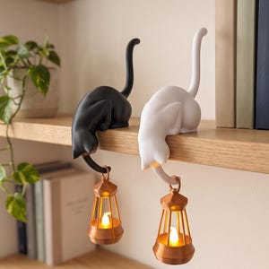 Puede incluir: Dos figuras de gato, una negra y otra blanca, están posadas en el borde de un estante. Cada gato tiene una cola larga y erguida, y sostiene una pequeña linterna de color cobre con una luz brillante desde su cola curvada hacia abajo.