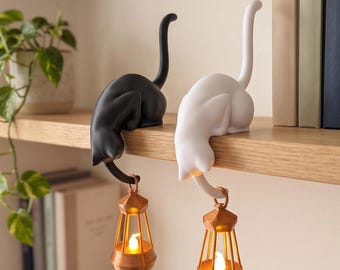 May include: Two cat figurines, one black and one white, are perched on the edge of a shelf. Each cat has a long, upright tail and holds a small, metallic copper-coloured lantern with a glowing light inside from its downward-curving tail.