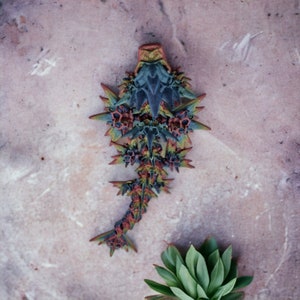 May include: A 3D printed model of a stylized creature with a blue, green, and red color scheme. The creature has a spiky, geometric design and is posed on a light brown surface.