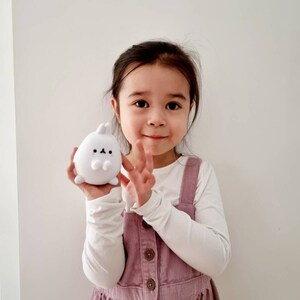K&ouml;nnte beinhalten: Ein junges M&auml;dchen tr&auml;gt ein wei&szlig;es Langarmshirt und ein rosafarbenes Latzkleid und h&auml;lt ein wei&szlig;es Pl&uuml;schkaninchen in der Hand. Das Kaninchen hat schwarze Augen und eine kleine schwarze Nase.