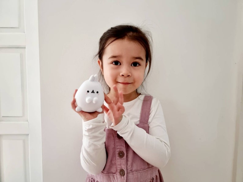 K&ouml;nnte beinhalten: Ein junges M&auml;dchen tr&auml;gt ein wei&szlig;es Langarmshirt und ein rosafarbenes Latzkleid und h&auml;lt ein wei&szlig;es Pl&uuml;schkaninchen in der Hand. Das Kaninchen hat schwarze Augen und eine kleine schwarze Nase.