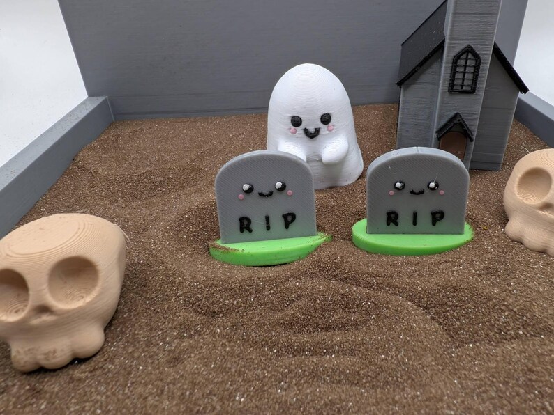 Pu&ograve; includere: Una scena di cimitero di Halloween con un fantasma bianco, due lapidi grigie con la scritta "RIP" su di esse e un teschio marrone. Le lapidi hanno dei volti carini disegnati su di esse.