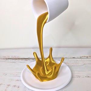 May include: A white ceramic cup appears to be pouring a stream of gold liquid onto a white plate. The liquid forms a splash shape. The background is a white wall and a light-colored wooden surface.
