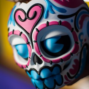 May include: A decorative skull with a white base, adorned with pink, blue, and black accents. The skull features large blue eyes and a detailed design, including a pink heart shape. The Day of the Dead-themed item is a vibrant piece of art.