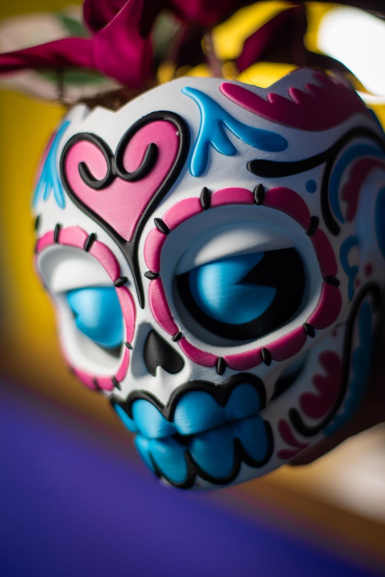 May include: A decorative skull with a white base, adorned with pink, blue, and black accents. The skull features large blue eyes and a detailed design, including a pink heart shape. The Day of the Dead-themed item is a vibrant piece of art.