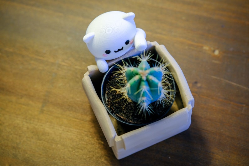 May include: A white ceramic figurine of a cat with a cute face, holding a small brown pot with a green cactus plant inside. The pot is sitting in a larger, light brown planter.