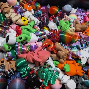Puede incluir: Una colección de juguetes articulados impresos en 3D, coloridos, que incluyen ranas, osos y otras criaturas. Los juguetes son de varios colores, incluyendo verde, rosa, naranja y patrones de arcoíris. Los juguetes están apilados.