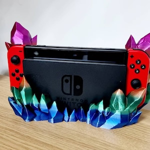 May include: A 3D printed stand for a Nintendo Switch console, featuring a crystal-like design with blue, green, and purple accents. The stand has a red and black Nintendo Switch console resting on it.