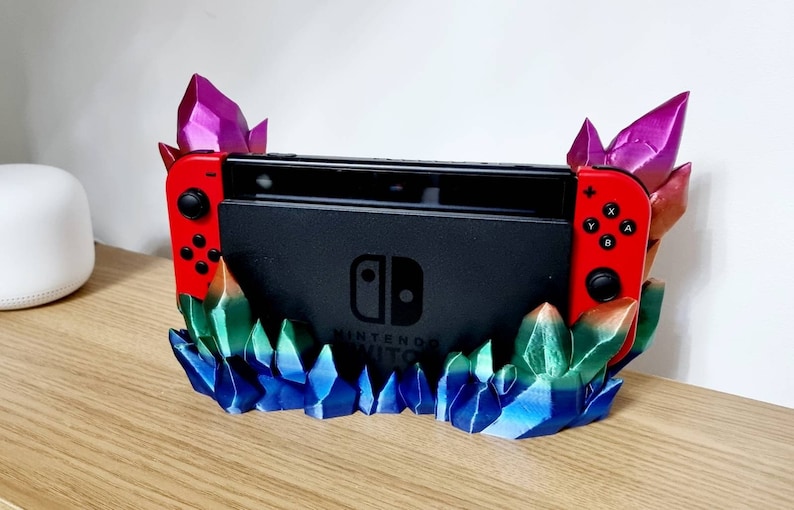 May include: A 3D printed stand for a Nintendo Switch console, featuring a crystal-like design with blue, green, and purple accents. The stand has a red and black Nintendo Switch console resting on it.