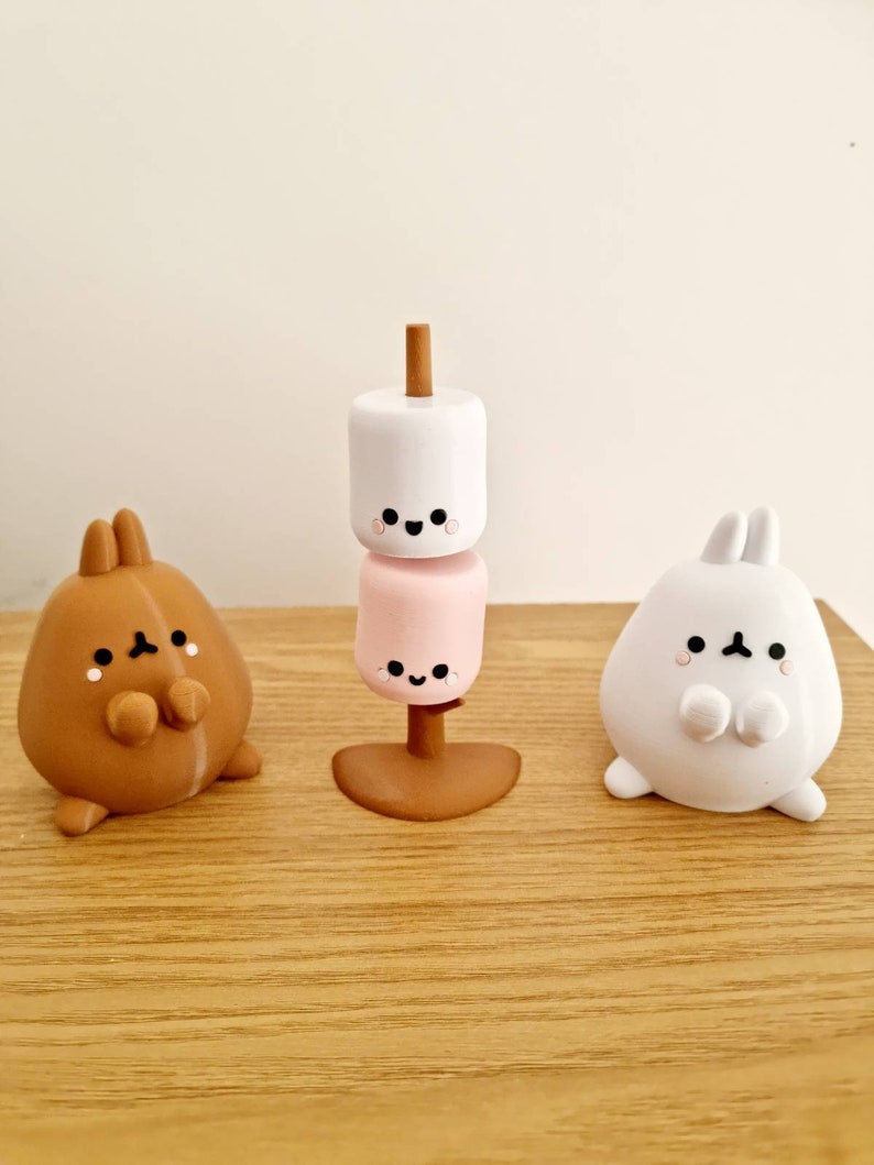 Op de afbeelding: Drie kleine, witte, bruine en roze keramische beeldjes van een konijn met een glimlachend gezicht. Het konijn links is bruin, het konijn rechts is wit en het konijn in het midden is een stapel van twee marshmallows op een stok.