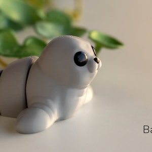 May include: A small, gray, baby seal figurine with black eyes. The figurine is lying on its side with its head turned to the right. The figurine is made of a smooth, plastic-like material.