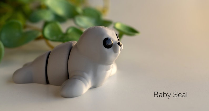 May include: A small, gray, baby seal figurine with black eyes. The figurine is lying on its side with its head turned to the right. The figurine is made of a smooth, plastic-like material.