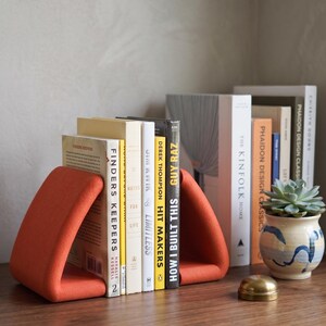 May include: A row of books held by orange triangular bookends. The books have various colored spines and titles, including "Finders Keepers" and "How I Built This". A small succulent in a blue and white pot sits nearby.