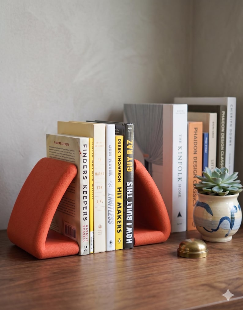 May include: A row of books held by orange triangular bookends. The books have various colored spines and titles, including "Finders Keepers" and "How I Built This". A small succulent in a blue and white pot sits nearby.