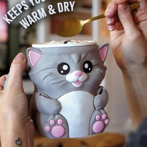 May include: A gray cat-shaped mug with pink accents, holding a golden spoon. The mug has a white interior and the text "KEEPS YOUR HANDS WARM & DRY". The mug's design includes large black eyes, a pink nose, and pink paw prints.