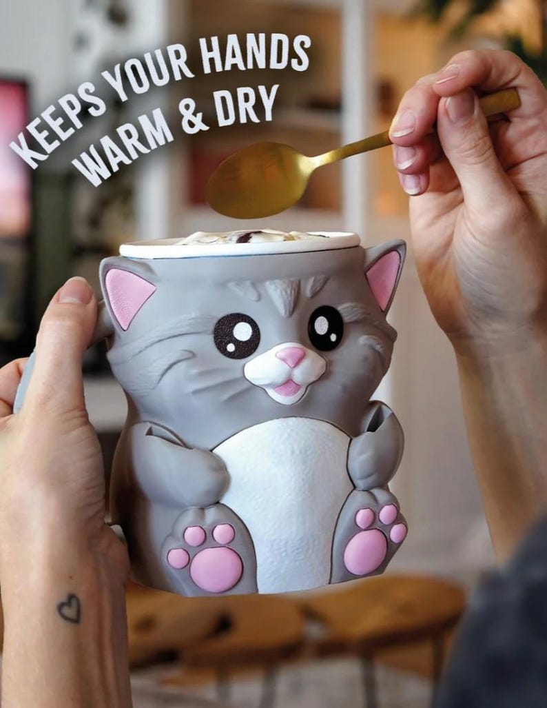 May include: A gray cat-shaped mug with pink accents, holding a golden spoon. The mug has a white interior and the text "KEEPS YOUR HANDS WARM & DRY". The mug's design includes large black eyes, a pink nose, and pink paw prints.