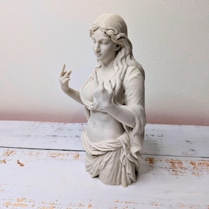May include: A matte off-white bust sculpture of a figure with long flowing hair, wearing draped fabric around the waist and chest. The figure is positioned with both hands raised, each displaying a middle finger gesture in a defiant pose.