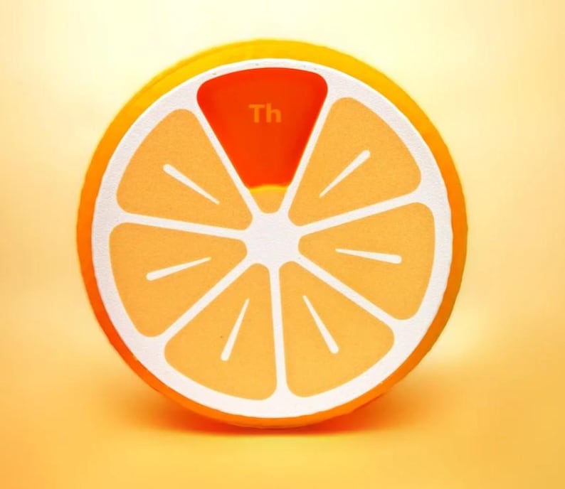 May include: A round, orange-colored object resembling a citrus fruit slice, with white segments and a bright orange rind. The top segment is a darker orange and has the letters "Th" printed on it.