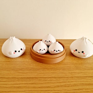 May include: Four white ceramic dumplings with cute faces, three are in a brown bamboo steamer, and one is on a wooden table.