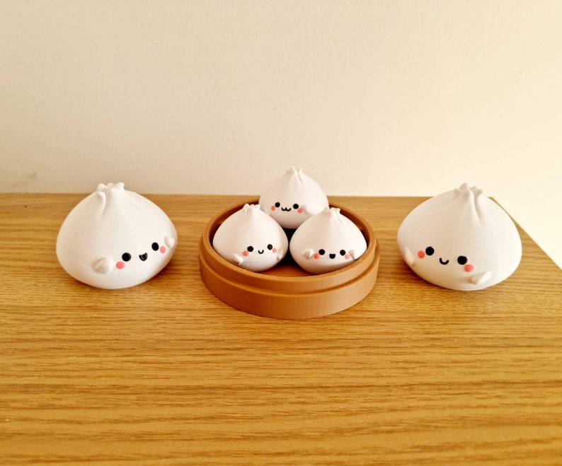 May include: Four white ceramic dumplings with cute faces, three are in a brown bamboo steamer, and one is on a wooden table.