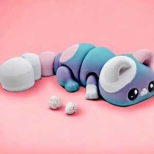 May include: A purple and blue cat figurine with white accents and large eyes. The cat is lying on its side with its tail curled up. There are two small white balls in front of the cat.