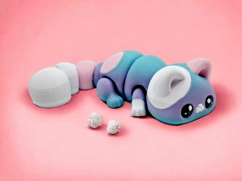 May include: A purple and blue cat figurine with white accents and large eyes. The cat is lying on its side with its tail curled up. There are two small white balls in front of the cat.
