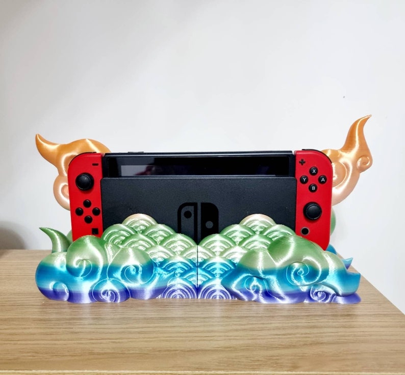 May include: A 3D printed stand for a Nintendo Switch console, featuring a colorful cloud design with gold accents. The stand has two horns on either side, resembling a mythical creature.