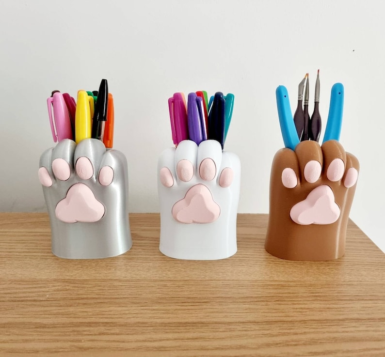 May include: Three paw-shaped desk organizers, one silver, one white, and one brown, each holding various pens and pencils.