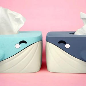 May include: Two whale-shaped tissue box holders. One is light blue, the other is dark blue. Both have white and cream-colored details, with a white tissue sticking out of the top. Each has a black curved eye and a white circle.