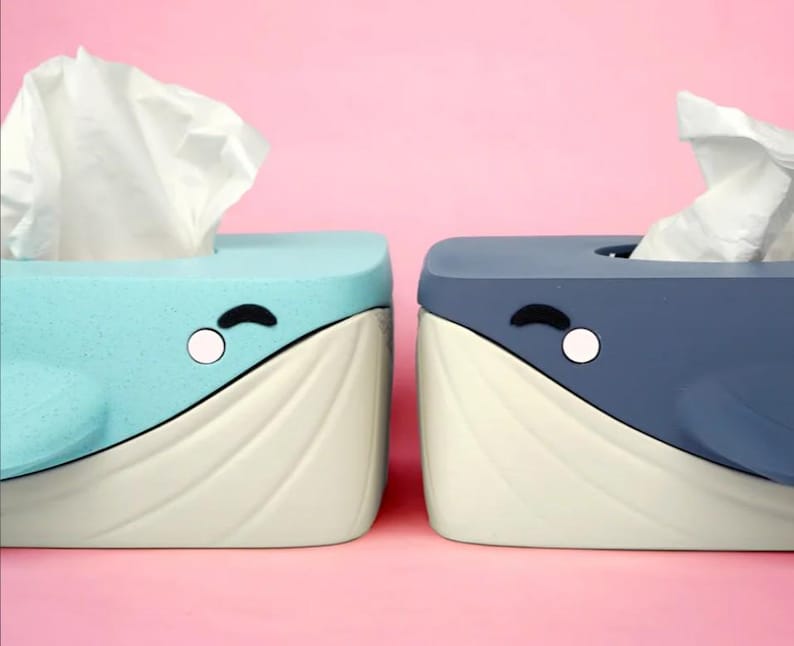 May include: Two whale-shaped tissue box holders. One is light blue, the other is dark blue. Both have white and cream-colored details, with a white tissue sticking out of the top. Each has a black curved eye and a white circle.